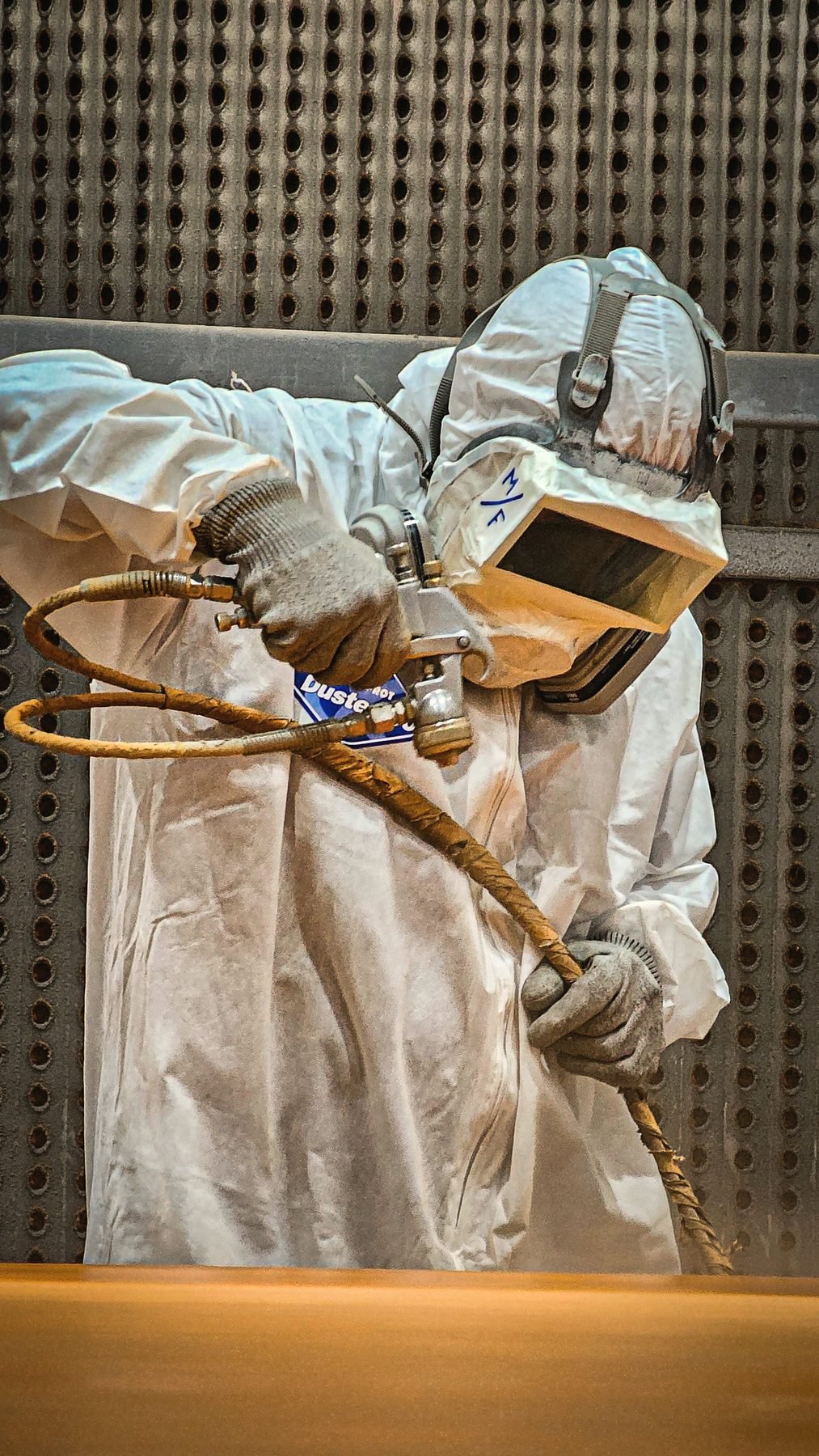 Operator in a hood and protective suit blasting a part against a perforated wall.